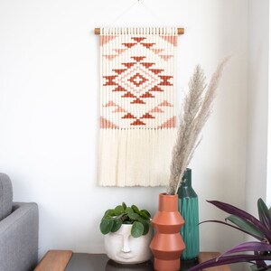 Könnte beinhalten: Ein Holz-Beistelltisch mit einer Glasplatte, auf dem ein Buch mit dem Titel "The Year of Knots" und eine Topfpflanze mit einem Gesichts-Topf stehen. Der Tisch steht neben einer Wand mit einem gewebten Wandbehang mit einem geometrischen Muster in Rosa- und Orangetönen.