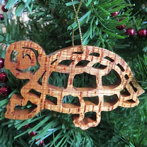 May include: Wooden turtle Christmas ornament with a smiling face and openwork shell design. The ornament is made of light brown wood and hangs from a gold string. The turtle is displayed on a green Christmas tree.