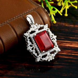 May include: A silver pendant featuring a large, red, rectangular gemstone set within a filigree frame. The frame is adorned with smaller, clear gemstones.