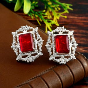 Könnte beinhalten: Ein Paar silberne Ohrringe mit einem roten quadratischen Edelstein, der in einem filigranen Design gefasst ist.