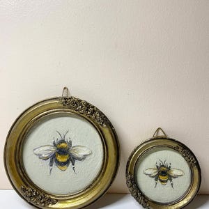Puede incluir: Dos pinturas redondas enmarcadas en oro con abejas. Las abejas est&aacute;n pintadas con rayas amarillas y negras, con alas blancas y grises. Los marcos tienen detalles ornamentados y un peque&ntilde;o lazo para colgar. El fondo es de color verde claro.