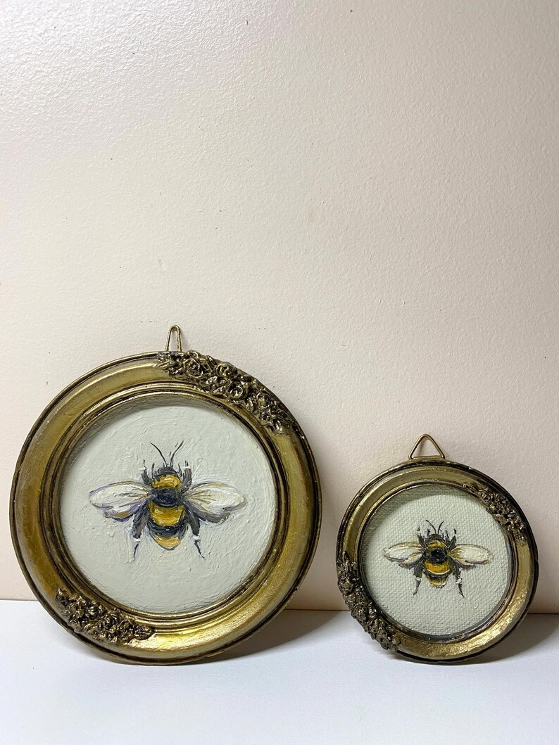 Puede incluir: Dos pinturas redondas enmarcadas en oro con abejas. Las abejas est&aacute;n pintadas con rayas amarillas y negras, con alas blancas y grises. Los marcos tienen detalles ornamentados y un peque&ntilde;o lazo para colgar. El fondo es de color verde claro.