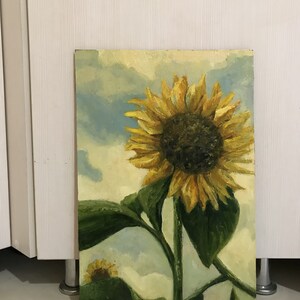 May include: A painting of a sunflower with yellow petals and a dark brown center. The sunflower is in the foreground of the painting, with a blue sky and white clouds in the background. The sunflower is painted in a realistic style, with visible brushstrokes.