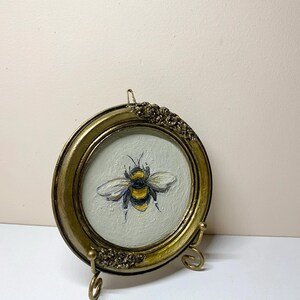 Puede incluir: Un peque&ntilde;o marco de fotos redondo de color oro antiguo con una pintura de una abeja. La abeja es amarilla, negra y blanca, sobre un fondo gris claro. El marco tiene detalles florales decorativos.