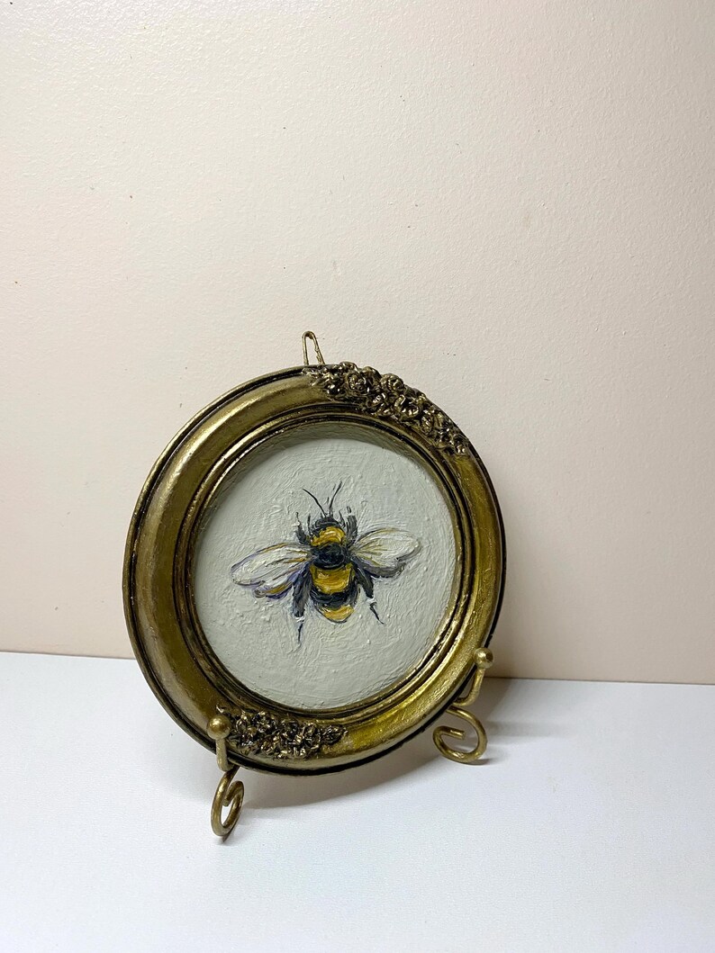 Puede incluir: Un peque&ntilde;o marco de fotos redondo de color oro antiguo con una pintura de una abeja. La abeja es amarilla, negra y blanca, sobre un fondo gris claro. El marco tiene detalles florales decorativos.
