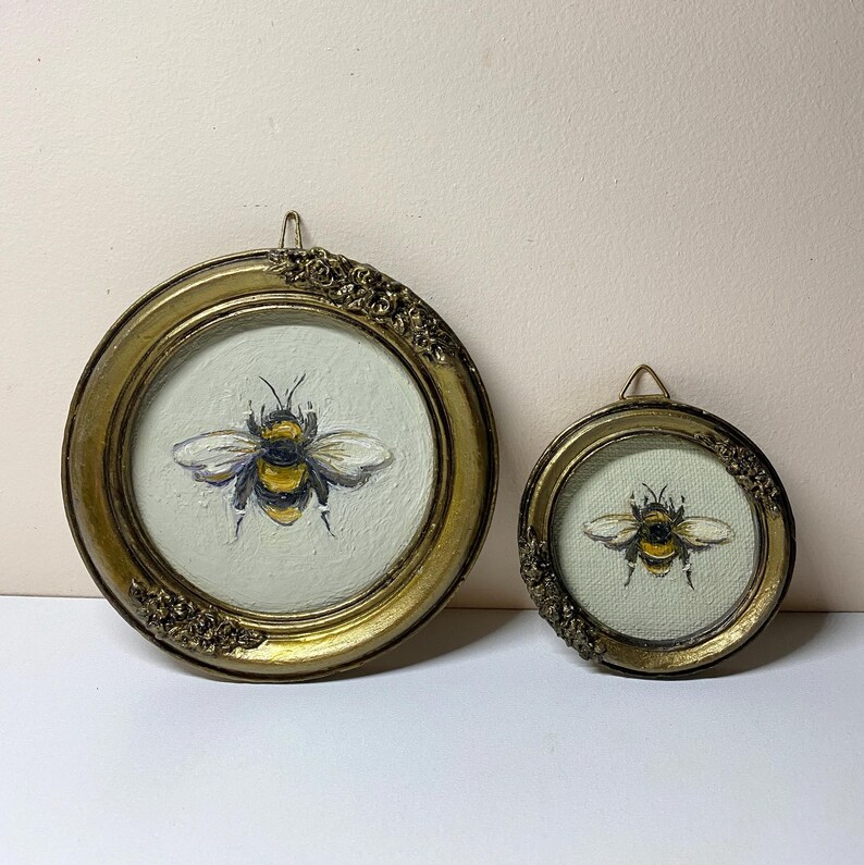 Puede incluir: Dos pinturas de abejas enmarcadas. Los marcos redondos son dorados con detalles ornamentados. Las pinturas de abejas presentan una abeja amarilla y negra con alas blancas sobre un fondo verde claro. Los marcos son de diferentes tama&ntilde;os.