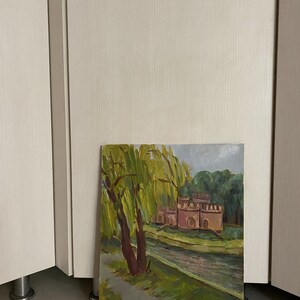 May include: A painting depicting a landscape with a river, a building, and a weeping willow tree. The building is a pale pink colour and has a few windows. The weeping willow tree is in the foreground and has green leaves. The river is in the background and is a light blue colour.