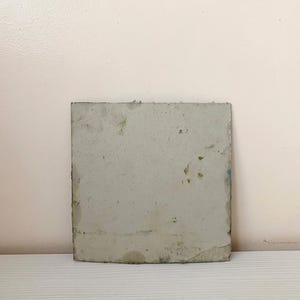 May include: A square, gray, ceramic tile with a slightly textured surface. The tile has a few small, green and blue spots on it.