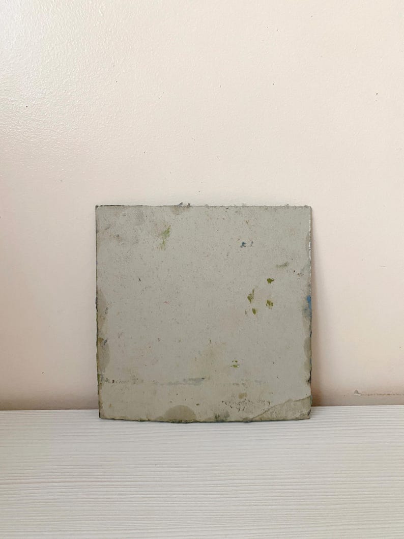 May include: A square, gray, ceramic tile with a slightly textured surface. The tile has a few small, green and blue spots on it.