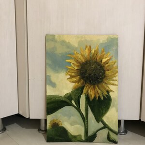 May include: A painting of a sunflower with yellow petals and a dark brown center. The sunflower is against a blue and white cloudy sky. The painting is on a canvas and has a wooden frame.