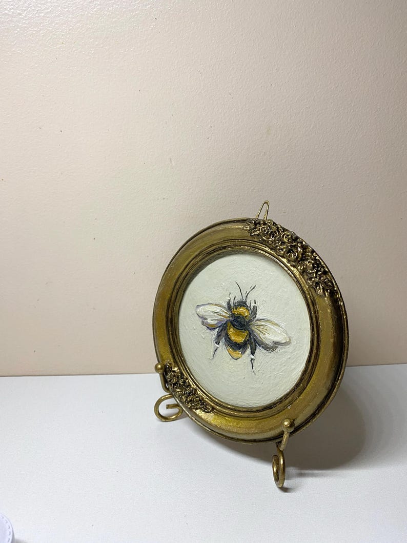 Puede incluir: Una peque&ntilde;a pintura redonda enmarcada en oro de una abeja. La abeja est&aacute; representada con rayas amarillas y negras y alas blancas. El marco tiene detalles ornamentados y se exhibe sobre un soporte dorado.