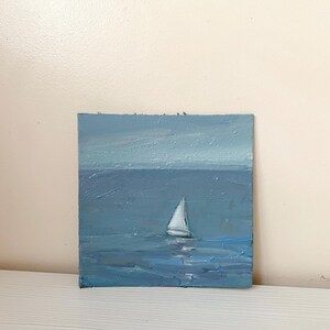 May include: A small oil painting of a sailboat on a blue sea. The sky is a light blue and the water is a darker blue. The sailboat is white and has a single sail.