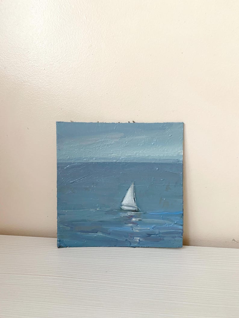 May include: A small oil painting of a sailboat on a blue sea. The sky is a light blue and the water is a darker blue. The sailboat is white and has a single sail.