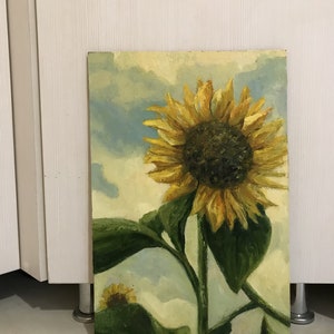 May include: A painting of a sunflower with yellow petals and a dark brown center. The sunflower is against a light blue and white cloudy sky. The sunflower is painted in a realistic style.