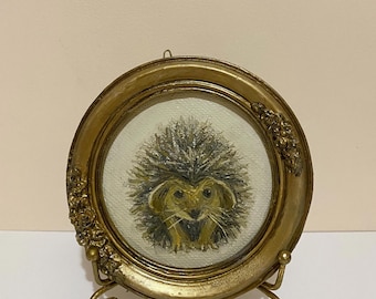 Puede incluir: Pequeña pintura redonda de un erizo en un marco dorado y ornamentado, presentada sobre un soporte decorativo. La obra de arte presenta un erizo con la espalda espinosa y una cara marrón claro. El marco mide unos 10 cm de diámetro.