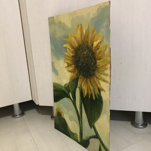May include: A painting of a sunflower with yellow petals and a dark brown center. The sunflower is against a background of blue and white clouds.