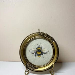 Puede incluir: Un marco redondo dorado antiguo con detalles florales en la parte superior e inferior. En el interior, una pintura de una abeja con rayas amarillas y negras y alas blancas. El marco se exhibe sobre un peque&ntilde;o caballete dorado.