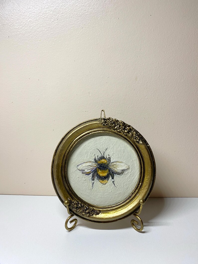 Puede incluir: Un marco redondo dorado antiguo con detalles florales en la parte superior e inferior. En el interior, una pintura de una abeja con rayas amarillas y negras y alas blancas. El marco se exhibe sobre un peque&ntilde;o caballete dorado.