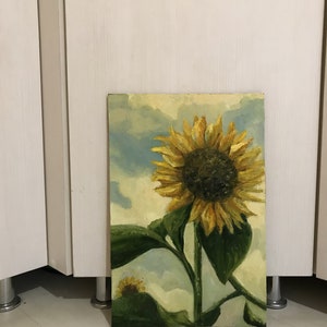 May include: A painting of a sunflower with a yellow center and yellow petals. The sunflower is against a blue and white cloudy sky. The sunflower is in a field of green leaves.