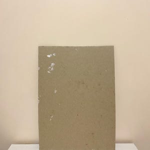May include: A plain brown cardboard sheet with white flecks on the surface. The sheet is sitting on a white surface.