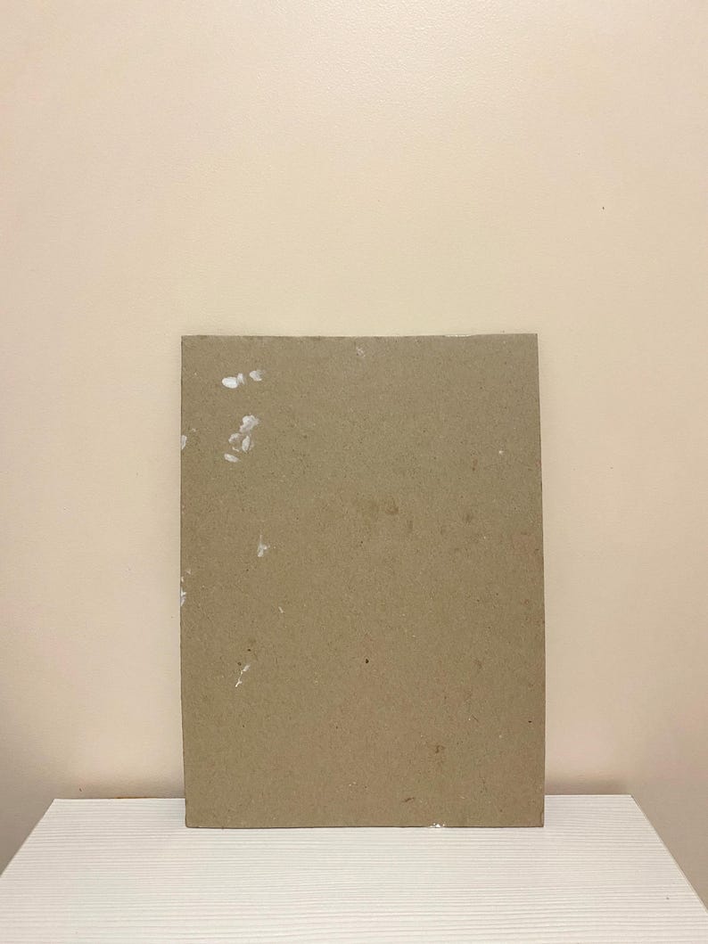 May include: A plain brown cardboard sheet with white flecks on the surface. The sheet is sitting on a white surface.