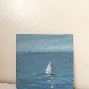 May include: A small oil painting of a sailboat on a blue sea with a light blue sky in the background.