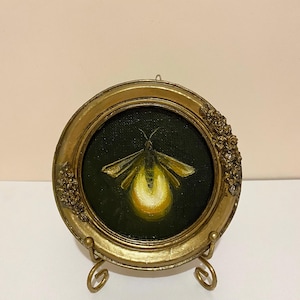 Puede incluir: Pequeña pintura redonda de una luciérnaga en un marco dorado ornamentado. La luciérnaga tiene un abdomen y alas de color amarillo brillante sobre un fondo verde oscuro. El marco está sostenido por un soporte dorado decorativo.