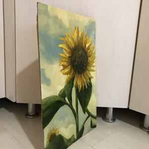 May include: A painting of a sunflower with yellow petals and a dark brown center. The sunflower is against a blue and white cloudy sky. The painting is on a wooden canvas.
