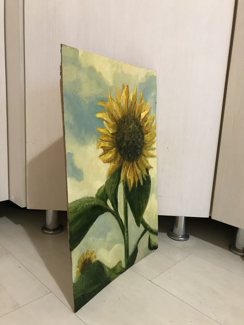 May include: A painting of a sunflower with yellow petals and a dark brown center. The sunflower is against a blue and white cloudy sky. The painting is on a wooden canvas.