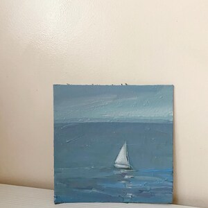 May include: A small oil painting of a sailboat on a blue sea with a light blue sky in the background.