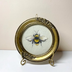 Puede incluir: Pintura redonda de una abeja en un marco dorado. La abeja est&aacute; pintada con detalles amarillos, negros y blancos. El marco tiene detalles florales y se muestra sobre un soporte dorado.