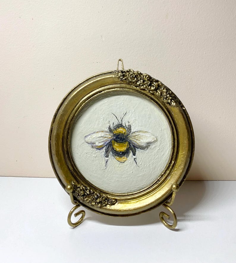 Puede incluir: Pintura redonda de una abeja en un marco dorado. La abeja est&aacute; pintada con detalles amarillos, negros y blancos. El marco tiene detalles florales y se muestra sobre un soporte dorado.