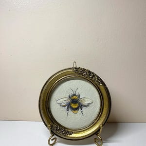 Puede incluir: Pintura redonda enmarcada en oro que representa una abeja. La abeja est&aacute; representada con rayas amarillas y negras y alas blancas. El marco tiene detalles florales decorativos y se exhibe sobre un peque&ntilde;o soporte dorado.
