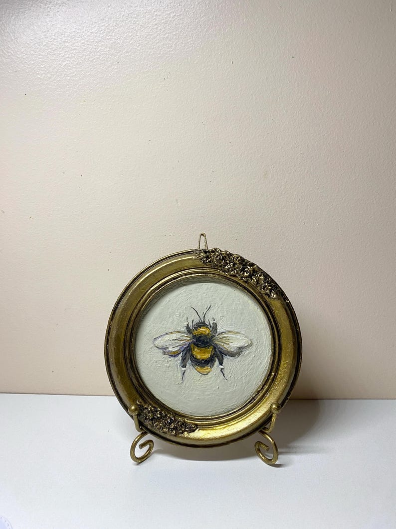 Puede incluir: Pintura redonda enmarcada en oro que representa una abeja. La abeja est&aacute; representada con rayas amarillas y negras y alas blancas. El marco tiene detalles florales decorativos y se exhibe sobre un peque&ntilde;o soporte dorado.