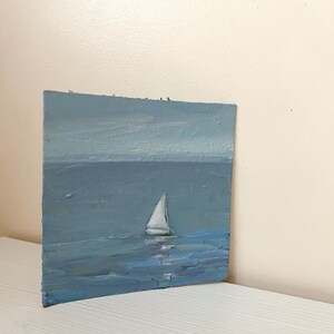 May include: A small painting of a sailboat on a blue sea with a light blue sky. The sailboat is white and has a single sail.
