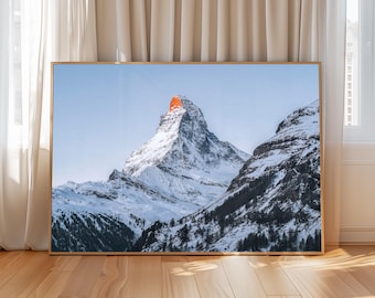 May include: A rectangular framed wall art print featuring a landscape photograph of the Matterhorn mountain peak covered in snow, with the summit illuminated by a bright orange sunrise glow against a clear pale blue sky and dark rocky slopes.