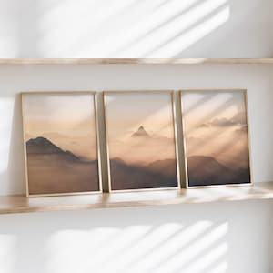 以下が含まれることがあります： 霧と日の出を背景にした山脈の額装されたプリントが3枚。プリントは金色の額縁に収められ、木製の棚に掛けられています。