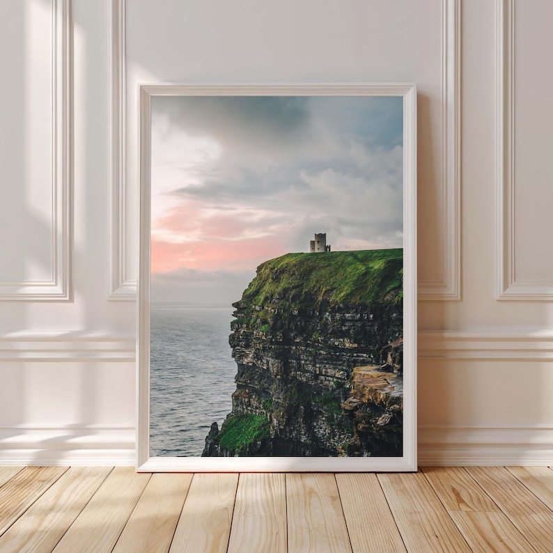 May include: A vertical framed wall art print depicting a stone castle perched atop a steep, dark cliff face covered in vibrant green grass. The scene overlooks a vast, calm ocean under a soft, pastel-colored sunset sky with light clouds.