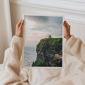May include: A framed vertical wall art print featuring a landscape photograph of a stone tower perched atop a steep, green cliff overlooking the ocean under a soft, cloudy sky with hints of pink light at sunset or sunrise.