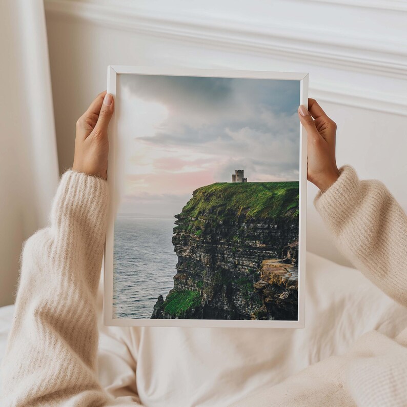 May include: A framed vertical wall art print featuring a landscape photograph of a stone tower perched atop a steep, green cliff overlooking the ocean under a soft, cloudy sky with hints of pink light at sunset or sunrise.