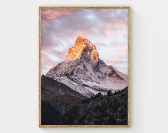 May include: A framed print featuring a majestic mountain peak illuminated by the warm glow of the setting sun. The snow-capped mountain is set against a backdrop of a pastel-colored sky, with a wooden frame.