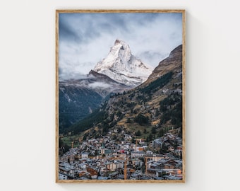 May include: A framed vertical wall art print featuring a snow-capped mountain peak towering over a dense alpine village. The landscape shows dark green pine forests on the slopes and a cluster of buildings with grey roofs under a cloudy sky.
