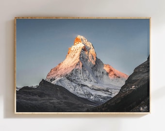 May include: A framed print of a snow-capped mountain peak, illuminated by the golden light of the setting or rising sun. The mountain is framed by a light-colored wooden frame, and the sky is a clear blue.