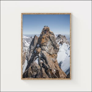 Może przedstawiać: Oprawione zdjęcie szczytu górskiego z ludźmi na szczycie. Góra jest skalista i brązowa, ze zboczami pokrytymi śniegiem i innymi szczytami w tle. Niebo jest czysto niebieskie.