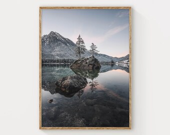 以下が含まれることがあります： 静かな山の湖の風景を描いた額装されたウォールアートプリント。穏やかな水面に雪を頂いた山脈が映り込み、中央には2本の常緑樹が立つ小さな岩の島が浮かんでいる様子が表現されています。