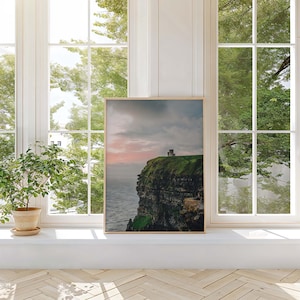 May include: A rectangular wall art print featuring a landscape photograph of a stone tower perched atop a steep, grassy cliff overlooking a dark ocean under a cloudy, soft pink and grey sky at dusk, framed in a light-colored wooden border.