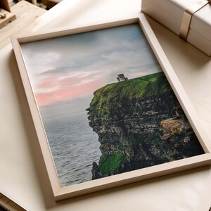 May include: A framed landscape photograph featuring a stone castle perched atop a steep, green-topped cliff overlooking the ocean. The sky displays soft pink and grey clouds at sunset, contrasting with the dark, layered rock face of the cliff.