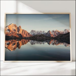 Könnte beinhalten: Gerahmter Landschaftsdruck mit einer Bergkette, die sich in einem ruhigen See spiegelt. Die Berge sind von warmem, goldenem Licht beleuchtet, das mit dem kühlen blauen Himmel und Wasser kontrastiert. Der Druck ist in einem hellen Holzrahmen.