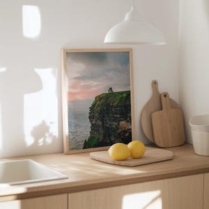 May include: A framed vertical wall art print featuring a landscape photograph of a green cliffside overlooking the ocean with a stone tower at the peak under a cloudy sunset sky, displayed on a wooden kitchen counter next to two lemons.