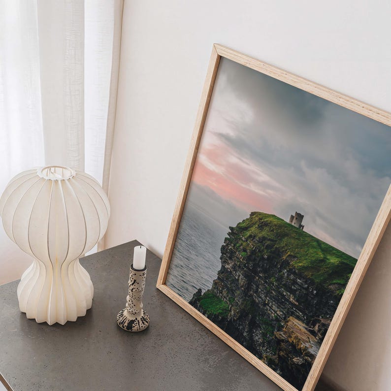 May include: A framed landscape photograph depicting a stone tower atop a green cliff overlooking the ocean at sunset, accompanied by a white pleated table lamp and a small speckled candle holder on a dark gray surface.